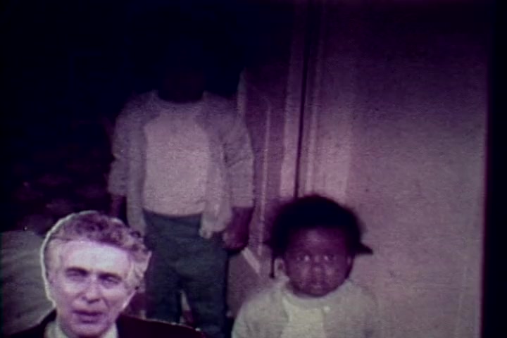 The still from 'Drug Abuse: Some Comments from the Morgue' features a somber scene, highlighting the emotional impact of drug abuse. In the foreground, a serious-looking man appears, likely Tillman Hahn from the Medical Examiner's office, who is discussing the consequences of drug use. In the background, two children are present, one obscured, adding a haunting element to the image. The muted colors and graininess typical of 16mm film emphasize the film's grim message and the toll of substance abuse on families.