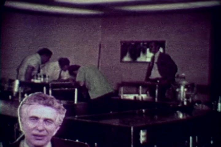 The still depicts a somber scene from the anti-drug film 'Drug Abuse: Some Comments from the Morgue.' In the foreground, a man, likely Tillman Hahn, is prominently featured, commenting on drug-related issues. Behind him, a group of four individuals in lab coats is gathered around a table, possibly examining evidence or conducting an investigation. The environment appears clinical and serious, reflecting the film's focus on the grim realities of drug abuse, likely emphasizing its consequences. The color tones are muted, characteristic of 1970s film quality.