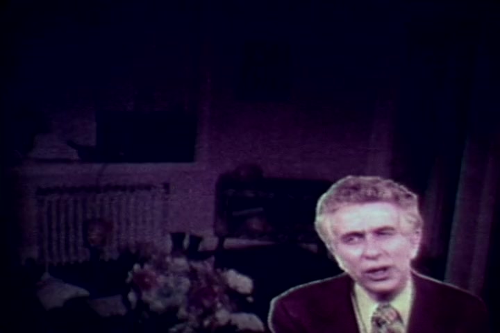 The still from 'Drug Abuse: Some Comments from the Morgue' features Tillman Hahn, identifiable by his distinctive suit and patterned tie, speaking directly to the camera. The background is dimly lit, hinting at a somber atmosphere typical of an anti-drug message. The setting appears to be a morgue or similar space, reinforcing the film's serious theme about the consequences of drug abuse. The overall tone is cautionary, focused on the grim realities associated with drug-related deaths.