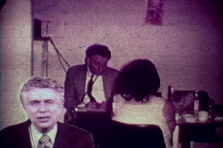 The still from 'Drug Abuse: Some Comments from the Morgue' features a split composition. In the foreground, a man in a suit, likely Tillman Hahn, addresses the camera directly. In the background, two individuals are seated at a table, one writing notes and the other facing away, suggesting a discussion or interview setting. The environment has muted colors typical of 1970s film, enhancing the serious tone of the anti-drug message. The overall mood reflects a sense of somber urgency about the consequences of drug abuse.