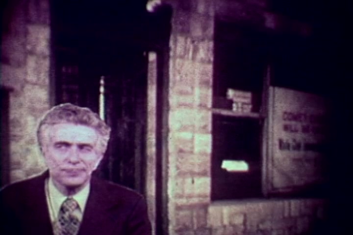 The still shows a somber-looking man, likely Tillman Hahn, in a suit with a tie, standing in front of a weathered brick building. The background includes a door and a window with a partially obscured sign, hinting at a grim context related to drug abuse. The color is muted, characteristic of the 1970s film quality, enhancing the serious tone of the anti-drug message.