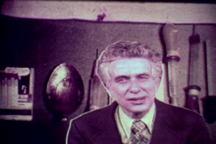 The still from 'Drug Abuse: Some comments from the morgue' features a man, likely Tillman Hahn, speaking directly to the camera. He appears serious and engaging, dressed in a suit with a patterned tie. Behind him, various tools and objects are visible, hinting at a medical or forensic context. The color appears faded, typical of 16mm prints from the 1970s, contributing to the film's historical and educational ambiance relating to the dangers of drug abuse.