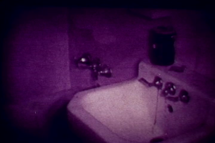 The still depicts a sink in a stark, subdued environment, characterized by a muted purple tint. Water appears to be flowing from the faucet, suggesting a sense of disarray or neglect. This imagery likely serves to underscore themes of drug abuse and its grim realities, as part of the anti-drug narrative conveyed by the film and its commentary by Tillman Hahn from the Medical Examiner's office. The setting evokes a somber atmosphere, fitting the film's serious subject matter.