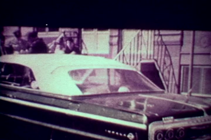 The still from 'Drug Abuse: Some comments from the morgue' features a classic car parked prominently in the foreground, likely from the 1960s. In the background, a group of individuals appears engaged in conversation outside a building, suggesting a neighborhood setting. The overall tone is gritty and reflective of the film's anti-drug message, characterized by its documentary style typical of the 1970s. The image captures a moment in urban life, invoking themes of societal issues related to drug abuse.