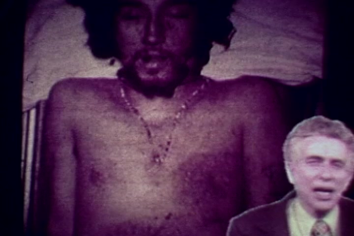 The still features a deceased individual lying on a morgue table, with a bare chest and visible chain necklace, evoking a somber tone. In the foreground, a man appears to be speaking, likely addressing the viewer about the consequences of drug abuse, typical of the film's intended anti-drug message from the 1970s. The color is muted, reflecting the serious nature of the subject matter, characteristic of educational films from that era.