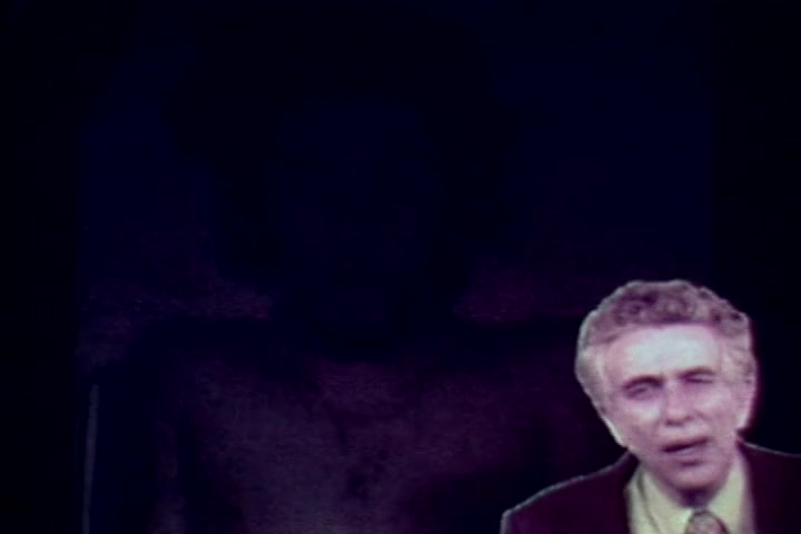 The still from 'Drug Abuse: Some Comments from the Morgue' features a dimly lit backdrop with a vague figure, likely representing a body. In the foreground, Tillman Hahn from the Medical Examiner's office is visibly speaking, appearing concerned as he addresses the audience. The overall tone is somber, reflecting the grim realities of drug abuse explored in the film. The image's graininess is characteristic of 16mm film, typical of that era.