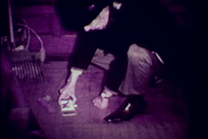 The still captures a somber scene from the anti-drug film 'Drug Abuse: Some comments from the morgue.' It features a figure, likely Tillman Hahn, crouched on the floor, inspecting a small box or container, which suggests a focus on the remnants of drug use. The image is tinged with a purple hue, characteristic of 16mm film from that era, conveying a sense of seriousness regarding the topic of drug abuse. The environment appears stark and clinical, reinforcing the film's grim message about the consequences of drug addiction.