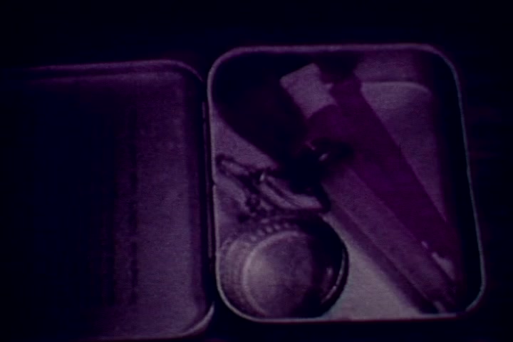 The still features an open tin box containing items associated with drug use, including a syringe and a small circular tin. The image has a purple tint, characteristic of the film's era. This visual reinforces the film's anti-drug message, emphasizing the stark reality of substance abuse.