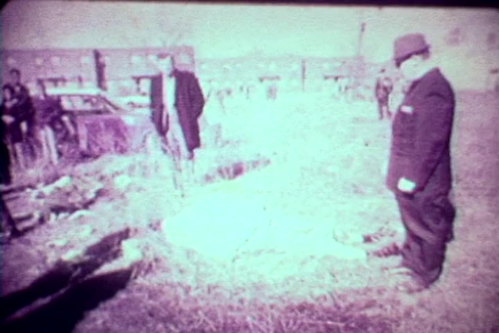 The still captures a somber scene from the 1970s anti-drug film, 'Drug Abuse: Some Comments from the Morgue.' It depicts two men, one in a suit and hat, examining a body on the ground amidst a desolate area. The background features a crowd of onlookers and dilapidated buildings, emphasizing the grim realities of drug abuse. The faded quality of the 16mm print adds to the stark and unsettling atmosphere of the moment.