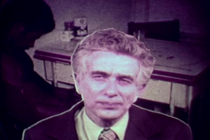 The still features Tillman Hahn, a medical examiner, prominently in the foreground, with a serious expression. The background includes a blurred figure, likely a subject of discussion, and a table with items that suggest a medical or investigative setting. The overall tone is somber, reflecting the film's anti-drug message from the 1970s, conveying the grim realities of drug abuse. The color palette is muted, characteristic of 16mm film, reinforcing the serious subject matter.