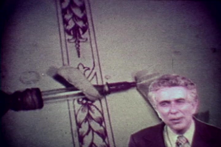 The still from 'Drug Abuse: Some comments from the morgue' features a speaker, likely Tillman Hahn, positioned in front of a backdrop that includes an ominous syringe and a wall decorated with ornate patterns. The overall tone suggests a serious examination of the consequences of drug abuse, aligning with the film's anti-drug message from the 1970s. The aesthetic reflects the era's educational filmmaking style, combining stark imagery with direct commentary.