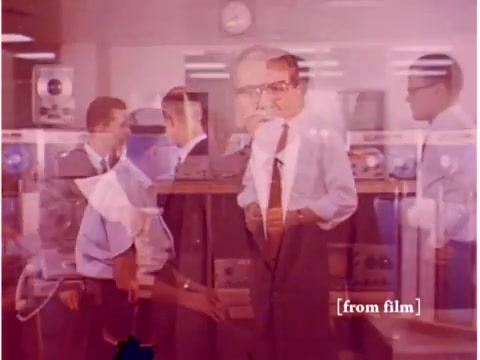 The still from the 1967 Bell System film depicts a group of engineers in a lab setting, surrounded by computers and equipment related to data transmission. The image is marked by a lab defect, resulting in a ghostly overlay effect where figures appear somewhat blurred and doubled. Key elements include a prominent clock in the background, various technical instruments likely related to acoustic modems or oscilloscopes, and individuals in professional attire engaged in discussion or monitoring equipment. The muted color palette adds a vintage feel, characteristic of the era's educational films.