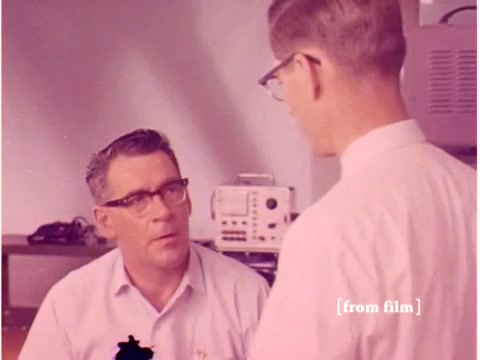 The still from the 1967 Bell System film depicts two engineers engaged in a discussion. One man, wearing glasses and a light-colored shirt, appears to be listening intently, while the other, also wearing glasses but in a white shirt, stands and seems to be explaining something. The background includes lab equipment, suggesting a technical environment focused on data transmission methods. The film is noted for its lab defects, indicating it may have visual anomalies or flaws.