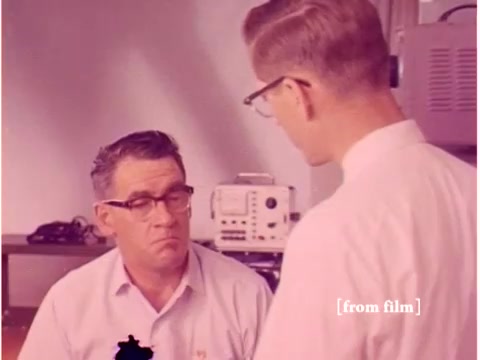 The still depicts two men in a laboratory setting, likely from the 1967 Bell System film focused on preventing data transmission errors. One man, wearing glasses and a light-colored shirt, appears to be listening intently while seated. The other, dressed in a white lab coat, is standing and engaging in conversation. The background features equipment characteristic of the era, such as computers and possibly an oscilloscope. The film is identified as a silent negative with some defects, affecting its overall clarity.