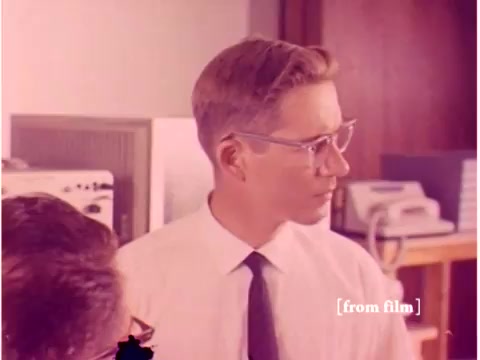 The still from 'Bell System - Preventing Data Transmission Errors' (1967) features two engineers in a lab setting. One engineer, wearing glasses and a tie, appears to be engaged in discussion or analysis. The background includes early computing equipment, indicative of the technological context of the era, such as computers and acoustic modems. The overall aesthetic reflects the 1960s, with a focus on data transmission and error prevention. The image is noted to be a lab defect, contributing to its unique vintage quality.