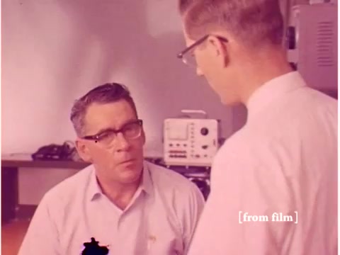 The still from 'Bell System - Preventing Data Transmission Errors - 1967' depicts two engineers engaged in conversation, likely discussing technical aspects of data transmission. The setting appears to be a laboratory, indicated by the presence of equipment like oscilloscopes in the background. The film quality shows some lab defects, adding a unique character to the visuals. This era's technology, including acoustic modems and paper tape, is central to the film's narrative on preventing transmission errors.