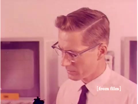 The still from the 1967 film 'Bell System - Preventing Data Transmission Errors' features a man in a white shirt and tie, wearing glasses, focused on a task. He appears to be an engineer or technician, possibly working with computer equipment or data transmission tools. The background suggests a laboratory or tech environment, typical for the era, with visible computer hardware and instrumentation. The colors are muted, reflecting the film's age and the likely processing defects inherent to the silent film format.