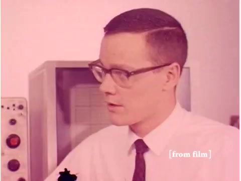 The still from the 1967 Bell System film features a young man wearing glasses and a tie, engaged in conversation. He is likely an engineer or technician, given the context of the film. Behind him, there is equipment that suggests a laboratory or technical environment. The overall aesthetic reflects the design and technology of the late 1960s, emphasizing the era's focus on telecommunications and data transmission. The film captures various elements like computers and oscilloscopes, showcasing technology from that time while having defects typical of older film stock.