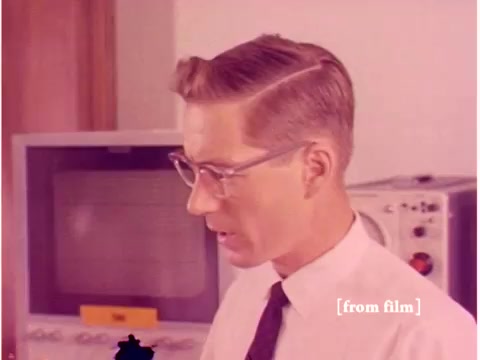 The still features a man with a neatly styled haircut, glasses, and a white shirt with a tie, suggesting a professional setting, likely in a scientific or technological environment. He appears to be focused on something out of frame, indicative of a thoughtful or analytical moment. In the background, there are vintage computing devices, including a monitor and various equipment typical of the 1960s era, which align with the film’s themes of data transmission and technology. The overall aesthetic has a muted color palette characteristic of the time period, with a hint of lab defects affecting the image.