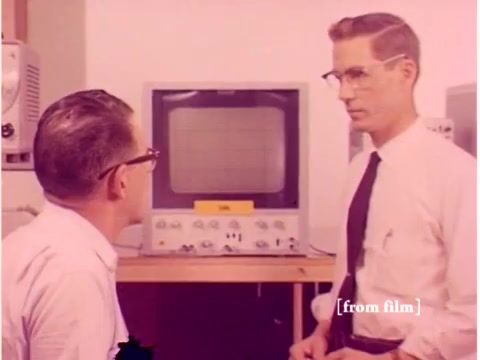 The still from 'Bell System - Preventing Data Transmission Errors' shows two engineers discussing in front of a computer monitor. The monitor displays a grid pattern, likely related to data transmission. One engineer, dressed in a light-colored shirt and glasses, observes the monitor intently, while the other, in a white shirt and tie, appears to be explaining something. The environment suggests a laboratory setting, with equipment hinting at the technological advancements of the 1960s. The film's silent nature emphasizes the visual interaction and the importance of the equipment being examined.