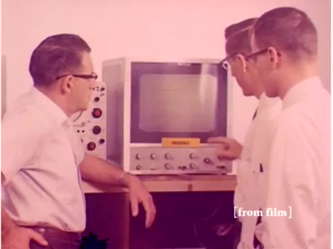 The still depicts three men, presumably engineers, gathered around a vintage monitor. The monitor displays a grid or oscilloscope-like pattern, indicative of data analysis or testing processes common in telecommunications. They're engaged in discussion, possibly troubleshooting or demonstrating equipment related to data transmission. The surrounding lab equipment suggests a focus on technology from the 1960s, emphasizing the era's approach to preventing data transmission errors. The overall atmosphere is technical and collaborative, reflecting the meticulous work involved in early computer and communication technology.