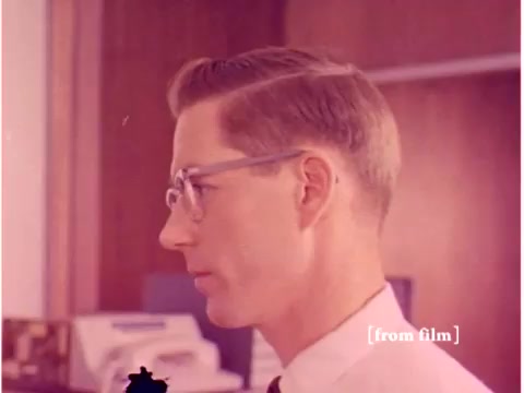 The still shows a profile view of a man wearing glasses and a suit, indicative of a professional setting, likely from the late 1960s. The background features blurred elements suggesting an office environment, possibly with equipment or papers associated with technology. This image, part of a silent film on data transmission errors by the Bell System, captures a moment of focus and engagement characteristic of engineering or technical work. As noted, the film has defects, which might affect the clarity or quality of the still.