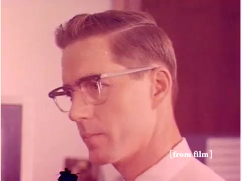 The still features a close-up of a man, likely an engineer or technician, prominently displayed with styled hair and wearing glasses. He has a focused expression, indicative of concentration on a task or discussion. The background suggests a laboratory or office environment typical of the 1960s, with hints of technology associated with data transmission. The color tones appear muted, possibly due to the film’s age or the lab defect mentioned. The overall context aligns with the themes of data processing and technological innovation during that era.