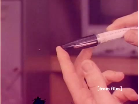 The still captures a close-up of a hand holding a pen, poised to touch the tip of a finger. The background features muted colors, likely due to the film defect, with a hint of equipment visible, suggestive of a lab or technical environment. The film's theme revolves around preventing data transmission errors in the context of the Bell System. The overall composition reflects a focus on the intersection of human interaction with technology, consistent with the film's subject matter.