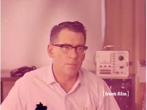 The still from the 1967 Bell System film features a man wearing glasses and a light-colored shirt, seated in what appears to be a lab or office environment. In the background, there is a piece of equipment, likely related to telecommunications or data transmission, suggesting a technical focus of the film. The image has a lab defect, which affects its clarity but adds to the vintage aesthetic. Overall, it captures a moment reflective of the era's technological advancements.