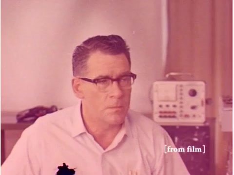 The still from 'Bell System - Preventing Data Transmission Errors - 1967' features a focused engineer with glasses sitting in a lab environment. He appears contemplative, possibly analyzing data or results related to the film's topic. In the background, there is a vintage piece of equipment, likely related to telecommunications or data transmission, consistent with the film's themes of oscilloscope footage and acoustic modems. The color tone suggests it’s from an older film stock, imparting a faded, historical feel to the scene. The film is noted to have lab defects, contributing to its unique character.