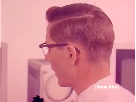 The still from 'Bell System - Preventing Data Transmission Errors' features a profile view of a man with a neat hairstyle wearing glasses, likely an engineer or technician from the 1960s. He is positioned in front of equipment that appears to be part of an early computing or communication setup, potentially involving oscilloscopes or acoustic modems. The film showcases the period's technology, emphasizing efforts to prevent data transmission errors. The image seems to suffer from a lab defect, affecting its clarity and visual quality.