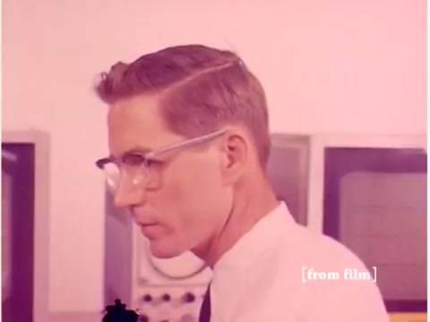 The still features a man with glasses and neatly styled hair, focused intently on the equipment around him. The background shows part of a laboratory setting with computing devices and oscilloscopes, reflecting the technological context of Bell System operations in the late 1960s. The lab defect has altered the colors, giving the image a pinkish tint, indicative of imaging issues rather than intentional color grading. The overall mood suggests a serious exploration of data transmission technologies during that era.