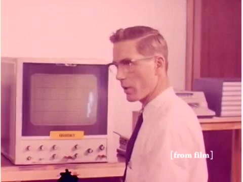 The still from the 1967 Bell System film shows a man in a white shirt and tie, wearing glasses, standing next to a large oscilloscope or monitor. The device is labeled 'FREQUENCY' and appears to be in a laboratory or office setting. The background includes stacks of what look like manuals or equipment, suggesting a technical environment focused on data transmission. The overall composition captures a moment in a historical context of technology and computer engineering, emphasizing an era when data transmission methods like acoustic modems were being explored.