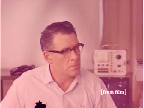 The still features a man with glasses, likely an engineer, engaged in thought or discussion, surrounded by equipment indicative of a lab setting. In the background, noticeable instruments suggest a focus on technology related to data transmission, aligning with the film's theme of preventing transmission errors. The overall aesthetic reflects the era's design and technological focus, typical of Bell System's work in the 1960s. The image quality appears to be influenced by lab defects, adding a unique character to the film.