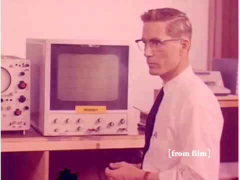 The still from 'Bell System - Preventing Data Transmission Errors - 1967' captures a focused engineer wearing glasses and a lab coat standing next to a large oscilloscope. The oscilloscope features a display screen with controls visible beneath it, hinting at its use for monitoring signals. In the foreground, additional electronic equipment is present, indicative of the technology used at the time. The overall scene reflects the period's dedication to developing and troubleshooting data transmission technology. The silent nature of the film emphasizes the visual aspects of the engineering process.