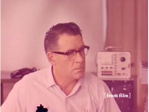 The still from 'Bell System - Preventing Data Transmission Errors - 1967' features an individual, likely an engineer, wearing glasses and a light-colored shirt. The background suggests a laboratory or technical setting, with equipment visible, including what seems to be an oscilloscope. The image has a slight color distortion, a characteristic of the lab defect noted in the film's description. Overall, it captures a moment of focus within a technical discussion or demonstration related to data transmission.