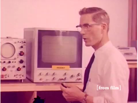 The still from the 1967 Bell System film features a man—likely an engineer—standing beside an oscilloscope and other laboratory equipment. He appears to be explaining or demonstrating something, as indicated by his gesturing hand. The color palette is muted, typical of vintage films from that era. The oscilloscope's screen is blank, and various knobs and dials are visible on the equipment behind him. The scene conveys a professional laboratory environment focused on data transmission and telecommunications technology. The lab defect suggests some visual distortion or quality issues due to its source.