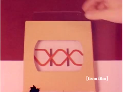 The still features a hand positioned to manipulate a piece of equipment, likely a model or schematic representation related to telecommunications. It showcases a cardboard frame displaying a design with intertwined lines reminiscent of waveforms or signals, indicative of data transmission concepts. The background is split into two colors, with a muted aesthetic typical of the era. This image aligns with the film's focus on preventing data transmission errors, highlighting the blend of visual aids and engineering practices from the late 1960s.