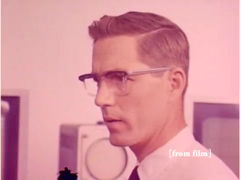 The still depicts a man with short, neatly combed hair and glasses, dressed in a white shirt with a tie. He appears to be focused and serious, suggesting a professional context. In the background, there are visible elements of a laboratory environment, including what looks like oscilloscopes and other computing equipment, typical of the Bell System's emphasis on technology and data transmission. The colors are muted, likely due to the film's age and laboratory defect, contributing to a retro aesthetic. Overall, the image reflects the mid-20th-century engineering and computing scene.