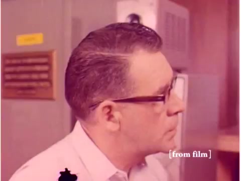 The still from the 1967 Bell System film shows a man with glasses and a neatly combed hairstyle, likely an engineer or technician, engaged in a setting indicative of a laboratory or technical workspace. The background features equipment and a plaque or sign, suggesting an environment focused on data transmission technology. The image’s color tones reflect the aesthetic of the era, with a slight defect affecting its clarity, typical of aging film stock. Overall, it captures a moment relevant to the technical explorations of the 1960s in telecommunications.