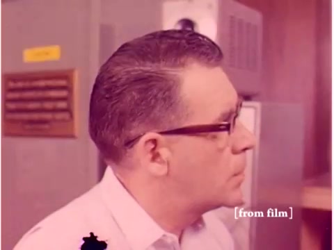 The still from the 1967 Bell System film shows a profile view of a man with short, neatly combed hair, wearing glasses and a light-colored shirt. In the background, there appears to be some lab equipment and a panel with buttons or switches, likely related to data transmission. The film captures the atmosphere of a technical environment prevalent in the late 1960s, reflecting the era's focus on communications technology. The image quality indicates it’s a silent negative from a defunct film lab, suggesting potential imperfections or lab defects in the film.