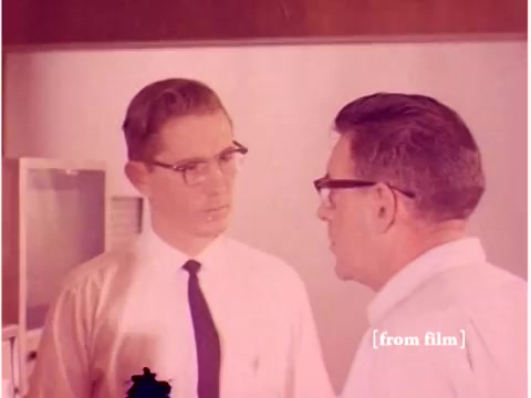 The still depicts two men engaged in a serious conversation, likely discussing technical issues related to data transmission. One man, wearing a tie and thin glasses, seems to be listening intently, while the other, also in glasses, appears to be explaining something. The background suggests a lab environment, possibly with computer equipment visible. The color saturation indicates it's a vintage 16mm film, and the overall scene conveys the atmosphere of a 1960s professional setting focused on technological analysis. The film's defect adds a slight imperfection, enhancing its vintage character.