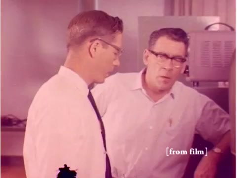 This still from 'Bell System - Preventing Data Transmission Errors' features two engineers engaged in discussion. The setting appears to be a laboratory or technical environment, indicated by the equipment in the background. The film has a muted color palette, typical of 1960s materials. This particular shot likely emphasizes the collaboration and expertise involved in preventing data transmission errors, reflecting the technological advancements of the era. The lab defect may contribute to a unique visual quality, capturing the essence of its time.