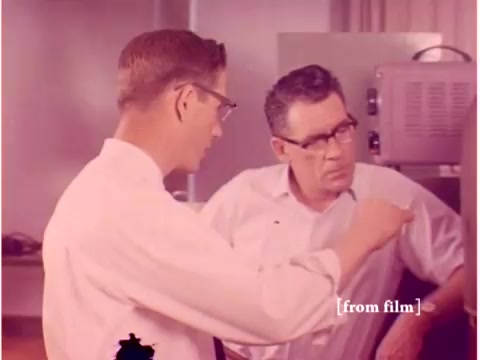 The still from the 1967 Bell System film features two engineers engaged in a discussion. One, wearing glasses and a white shirt, gestures towards an unseen object, likely relating to their work with data transmission. The second engineer, also in a professional outfit, appears attentive and is focused on the conversation. The background suggests a lab environment with equipment typical of the era, such as machines related to computing or data processing. The scene encapsulates a collaborative atmosphere in an engineering context, indicative of the era's technological advancements. The overall quality and colors reflect the film's age and the lab defect it came from.