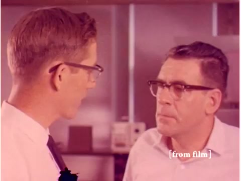 The still features two men engaged in a serious conversation, likely discussing technical matters related to data transmission or engineering. Both are wearing glasses, and their expressions suggest a focus on the topic at hand. The background hints at a laboratory or office setting, typical for the Bell System films of the era, aimed at illustrating technological processes. The image's color quality and the overall aesthetic reflect the visual style of the 1960s, while the film's status as a lab defect implies a lack of clarity or quality issues.