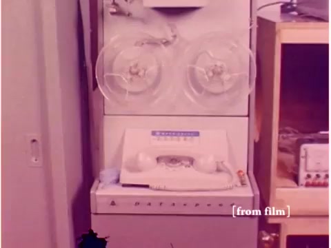 The still from the 1967 Bell System film showcases an early data processing machine. It features a tape drive system with two large reels, indicative of the technology of the time. Below the reels is a console with buttons and a telephone, possibly an acoustic modem setup. The background includes additional lab equipment, highlighting a technical environment focused on data transmission. The film's silent and defect status is noted, suggesting some visual imperfections. Overall, it captures a moment in the evolution of data communication technology.
