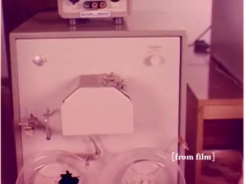 The still from the silent film 'Bell System - Preventing Data Transmission Errors - 1967' features a piece of laboratory equipment, likely related to data transmission or signal processing. The device has a prominent, boxy structure with valves or connectors on the front and appears to be connected to other machinery. The setting suggests a technical environment, typical of the time period, with wooden furniture visible in the background. The overall aesthetic reflects mid-20th century industrial design.