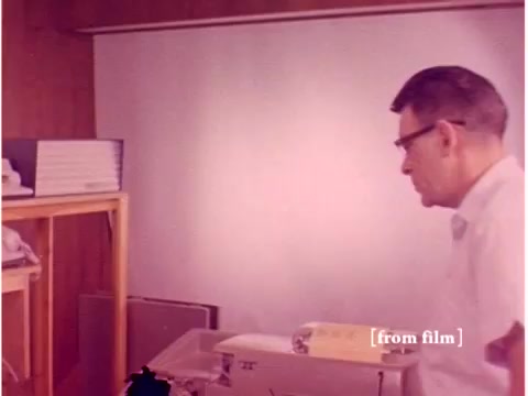 The still from 'Bell System - Preventing Data Transmission Errors' (1967) depicts an engineer in a lab setting, likely working with early computer technology. The environment features wooden paneling, a desk with electronic equipment, and stacks of materials, reflective of the era's lab aesthetics. The film appears to have some defects, affecting the image quality. Overall, it captures a snapshot of the technological experimentation and innovation focused on data transmission.