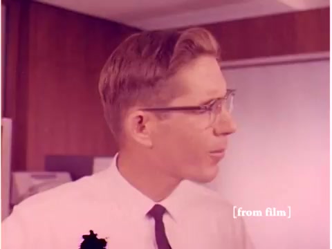 The still depicts a young man with glasses, possibly an engineer, looking off to the side in a professional setting. His hairstyle and attire suggest the 1960s era, typical of individuals in technical fields at that time. The background includes indistinct office elements, hinting at a laboratory or workspace where technological developments, like computers and modem technologies, are being discussed or demonstrated. The image likely contributes to the film’s theme of data transmission and error prevention in telecommunications. The overall quality seems affected by lab defects, potentially distorting colors or clarity.