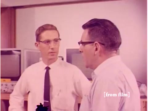 The still features two engineers engaged in a conversation, dressed in formal attire typical of the 1960s, with glasses. The background includes old computer monitors and lab equipment, indicative of a technology-focused setting. The overall color tone appears slightly faded, characteristic of a lab defect in the film. The context suggests a discussion about preventing data transmission errors, reinforcing the film's subject matter.